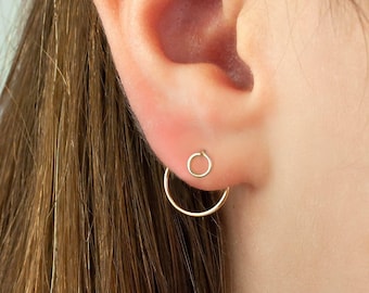 May include: A pair of gold-toned earrings consisting of a small circular stud worn in the upper ear lobe and a larger, thin circular hoop earring positioned below it, creating a layered, minimalist appearance on the ear.