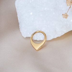May include: A gold-colored septum ring with a teardrop-shaped design. The ring has a smooth, polished surface and a hinged closure. The jewelry is set against a white stone and a neutral-toned background.