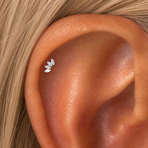 Puede incluir: Un pendiente de cartílago en tono plateado con tres piedras transparentes de corte marquesa dispuestas en un racimo curvo con forma de hoja. La joya está situada en la zona superior del cartílago de una oreja, mostrando un engaste metálico.