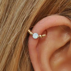 May include: A gold-colored hoop earring with a small, round, iridescent opal stone. The earring is designed with a coiled wire and small gold beads. The earring is worn in the ear cartilage.