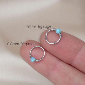 May include: Two silver-toned hoop rings, each featuring a small, round, light blue opal-like bead secured by wire wrapping. The larger ring is labeled 1mm-18gauge, and the smaller ring is labeled 0.8mm-20gauge, indicating their respective thicknesses.