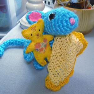 May include: A handmade crochet toy set featuring a blue mouse with pink ears and nose, holding a yellow teddy bear with a heart-shaped patch. A yellow and orange blanket is draped over the mouse. The toys are on a gray surface.