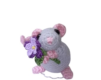 May include: A small, light gray crocheted mouse figurine with pink ears, nose, and feet. The mouse has black bead eyes and is holding a crocheted purple flower with a yellow center and green leaves in its front paws.