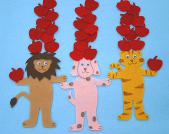 May include: Three felt animal figures, a lion, a dog, and a tiger, each with a stack of red felt apples on their heads. The lion is holding a red apple in its paw. The tiger is holding a red apple in its paw. The dog is holding a red apple in its paw.