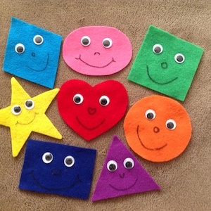 May include: A collection of colorful felt shapes, including a blue square, pink oval, green square, yellow star, red heart, orange circle, blue rectangle, and purple triangle. Each shape has googly eyes and a smiling mouth.