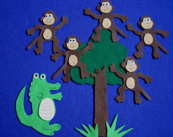 May include: A felt board scene featuring six brown monkeys with tan bellies and faces, positioned on a green tree with a brown trunk. A green crocodile is at the bottom. The background is a solid blue.