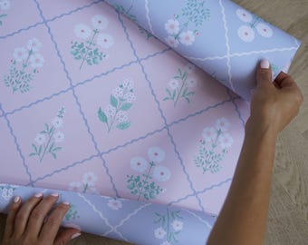 Può includere: Carta da regalo a motivi floreali nei toni del rosa e del blu. Il design presenta fiori bianchi e foglie verdi disposti a griglia. La carta si sta srotolando, rivelando il motivo.