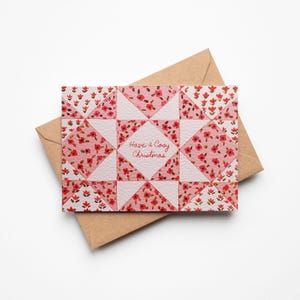 以下が含まれることがあります： ピンクと白の花柄キルト模様のクリスマスカード。中央には「Have a Cozy Christmas」の文字。茶色の封筒の上に置かれ、お祝いのメッセージを送るのに最適です。