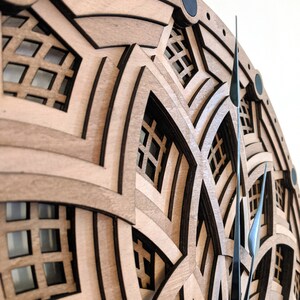 Op de afbeelding: Een gelaagde houten wandklok met een geometrisch ontwerp. De klok heeft lichtbruin hout met donkerbruine accenten en zwarte cirkelvormige details. De wijzers van de klok zijn donker metaalkleurig.
