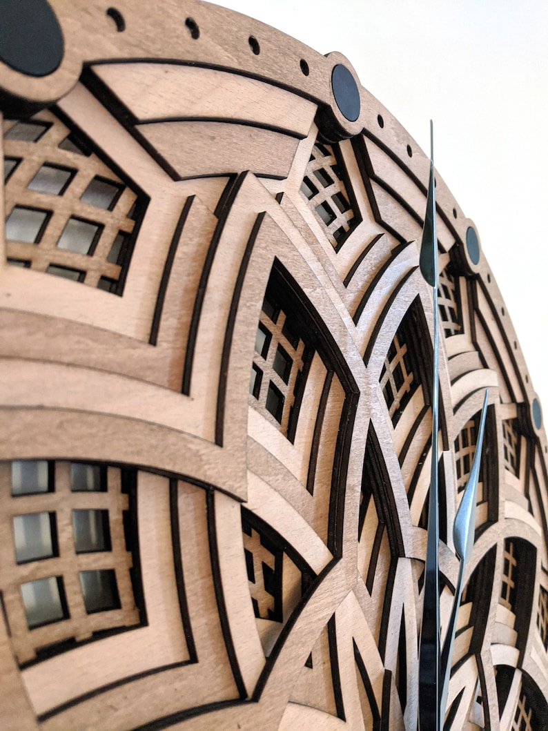Op de afbeelding: Een gelaagde houten wandklok met een geometrisch ontwerp. De klok heeft lichtbruin hout met donkerbruine accenten en zwarte cirkelvormige details. De wijzers van de klok zijn donker metaalkleurig.