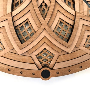 Op de afbeelding: Een gelaagde houten klok met een geometrisch ontwerp. De klok heeft een lichtbruin en donkerbruin kleurenschema, met ingewikkelde uitgesneden patronen. De wijzers zijn rechtsboven zichtbaar. De klok heeft een zwart midden en een cirkelvormig frame.