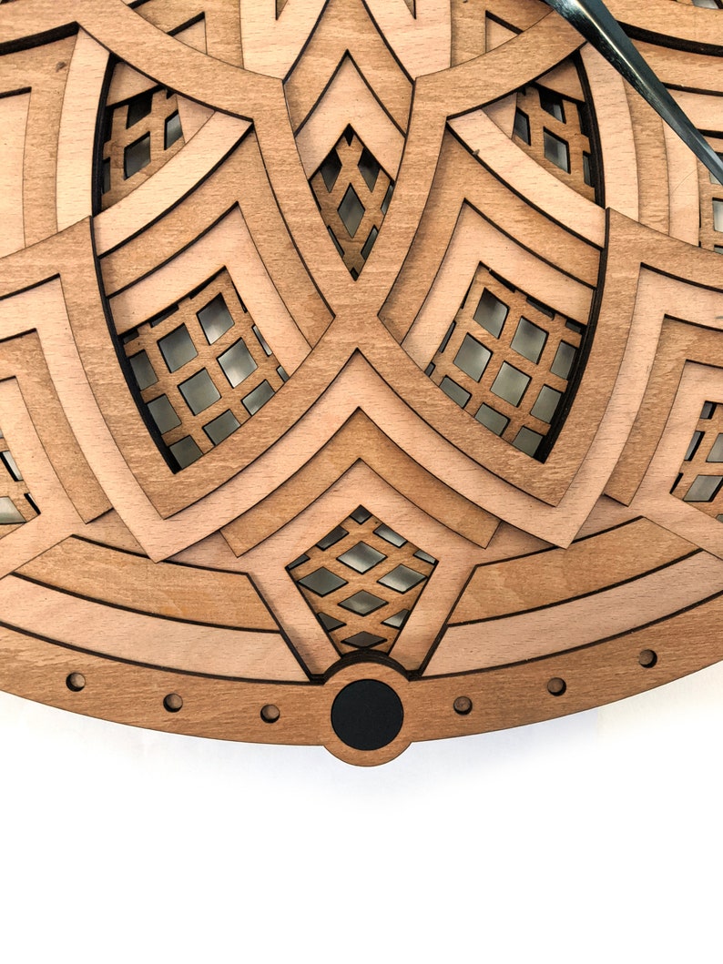 Op de afbeelding: Een gelaagde houten klok met een geometrisch ontwerp. De klok heeft een lichtbruin en donkerbruin kleurenschema, met ingewikkelde uitgesneden patronen. De wijzers zijn rechtsboven zichtbaar. De klok heeft een zwart midden en een cirkelvormig frame.