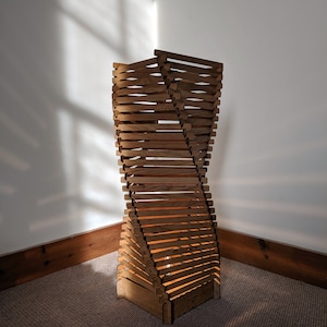 May include: A wooden floor lamp with a unique, twisted design. The lamp is constructed from horizontal wooden slats, creating an open, spiral shape. The warm light from within illuminates the wooden structure, casting shadows on the wall.