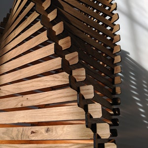 May include: A wooden sculpture made of stacked, angled pieces of wood. The wood is a light brown color and the sculpture has a spiral shape.