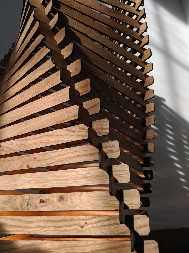 May include: A wooden sculpture made of stacked, angled pieces of wood. The wood is a light brown color and the sculpture has a spiral shape.