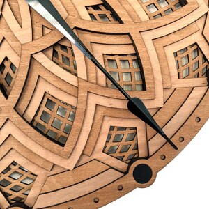 Op de afbeelding: Close-up van een houten klok met een gelaagd ontwerp. De klok heeft een geometrisch patroon met vierkante uitsparingen en zwarte wijzers. Het hout heeft een natuurlijke, lichtbruine kleur en de wijzers zijn zwart.