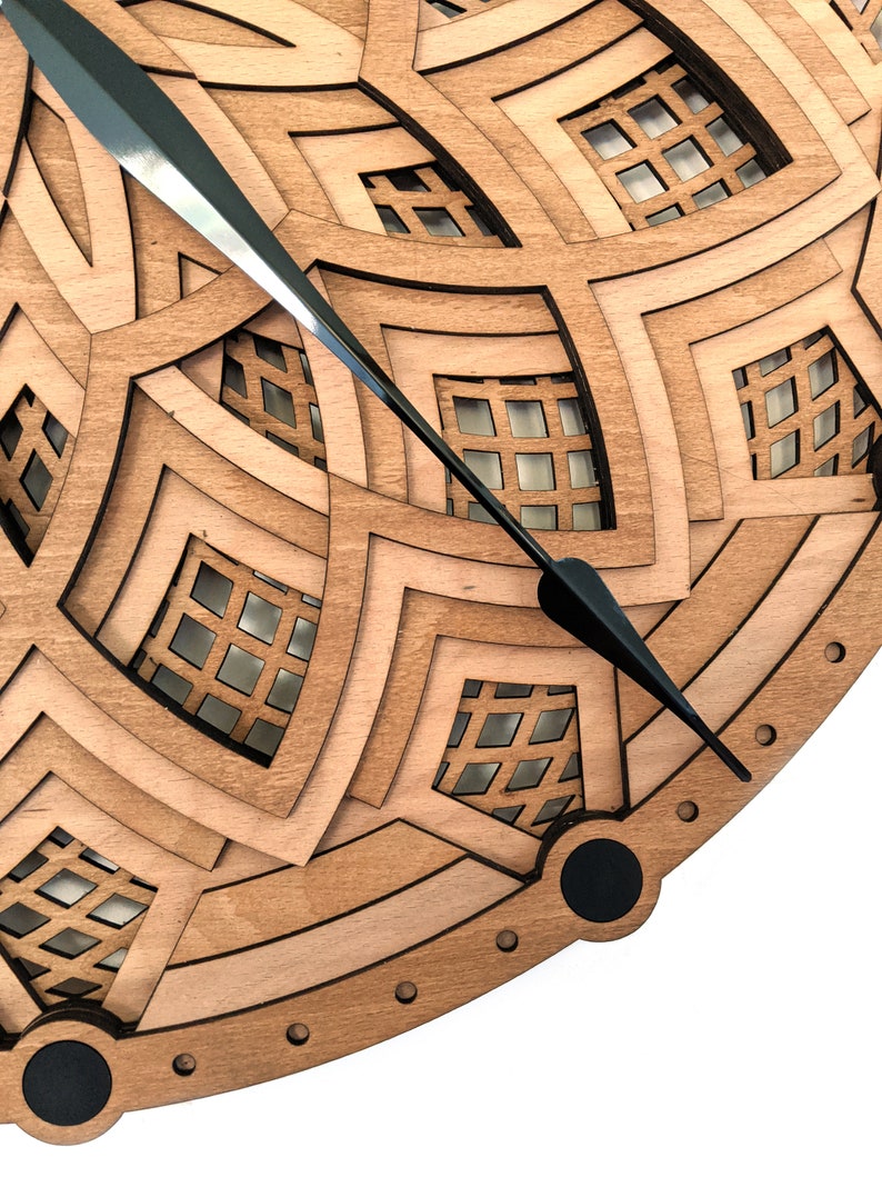 Op de afbeelding: Close-up van een houten klok met een gelaagd ontwerp. De klok heeft een geometrisch patroon met vierkante uitsparingen en zwarte wijzers. Het hout heeft een natuurlijke, lichtbruine kleur en de wijzers zijn zwart.