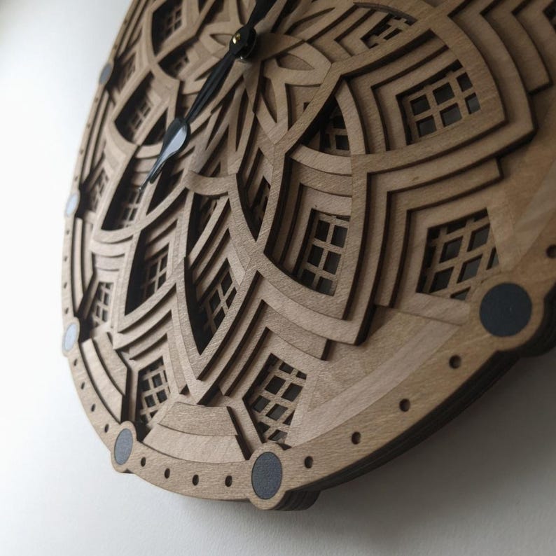 Op de afbeelding: Een houten wandklok met een gelaagd, geometrisch ontwerp. De klok heeft een lichtbruine afwerking met ingewikkelde uitsparingen en zwarte wijzers. Het ontwerp bevat herhalende patronen en kleine, vierkante raamachtige vormen. De diameter van de klok is ongeveer 30 cm.