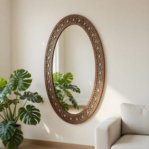 May include: Oval-shaped brown wooden mirror with a decorative, openwork border. The mirror is mounted on a light-colored wall. A potted plant with large green leaves is visible in the foreground, and a white sofa is in the background.