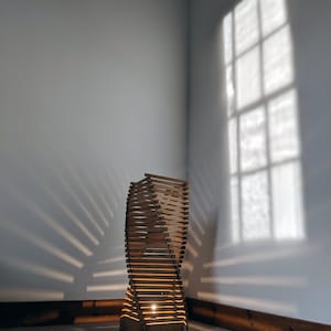 May include: A wooden floor lamp with a twisted design, illuminated by a light bulb. The lamp casts a shadow on the wall behind it.