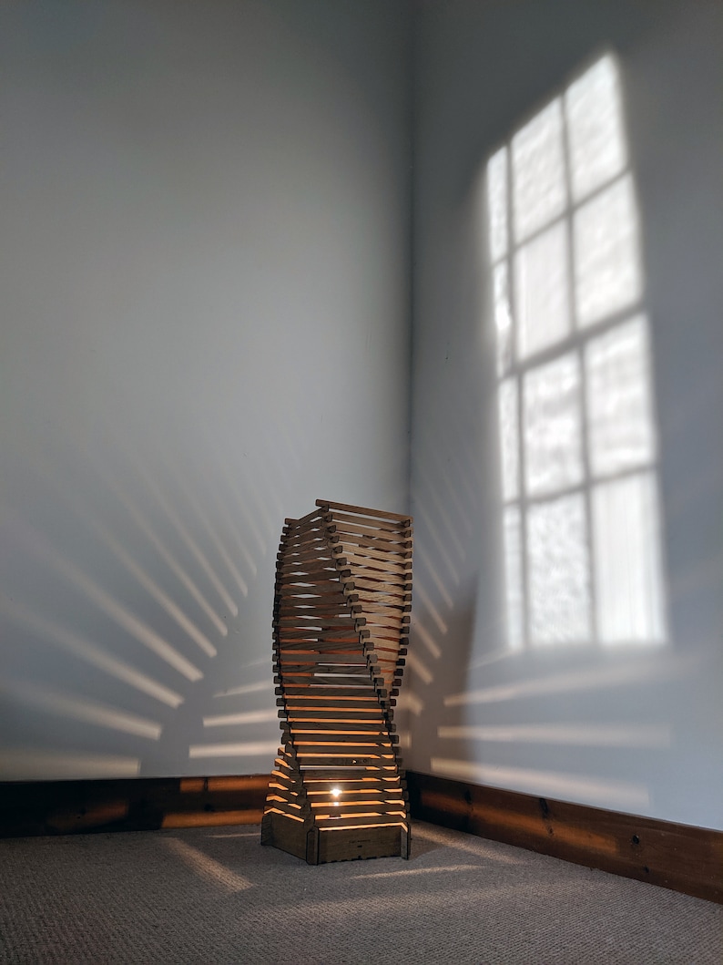 May include: A wooden floor lamp with a twisted design, illuminated by a light bulb. The lamp casts a shadow on the wall behind it.