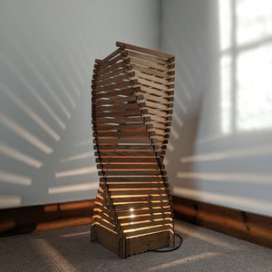 May include: A wooden floor lamp with a unique twisted design. The lamp is made of thin wooden slats and has a warm, natural finish. The lamp is lit and casting a warm glow on the floor.
