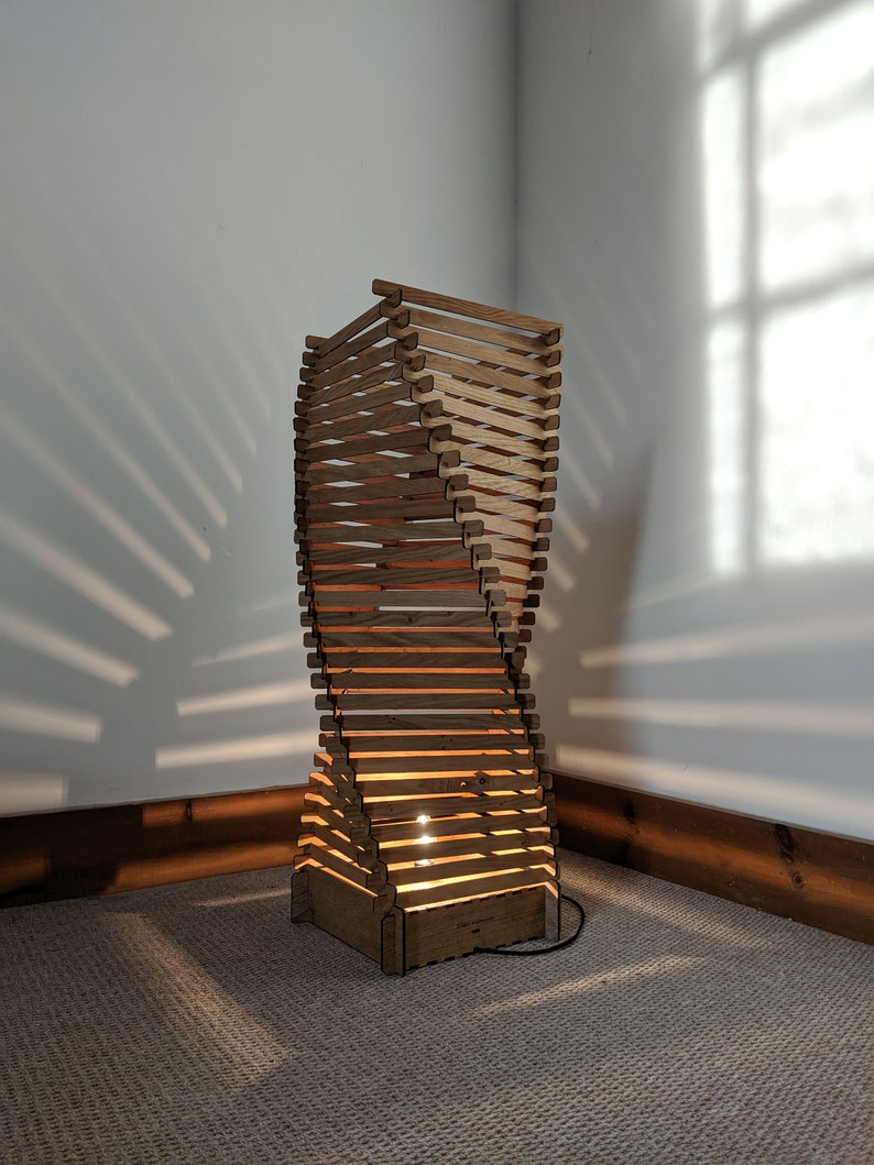 May include: A wooden floor lamp with a unique twisted design. The lamp is made of thin wooden slats and has a warm, natural finish. The lamp is lit and casting a warm glow on the floor.