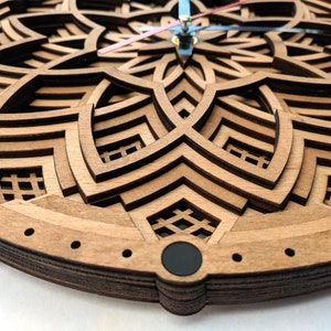 Op de afbeelding: Een houten wandklok met een gelaagd, geometrisch ontwerp. De klok heeft een lichtbruine kleur met ingewikkelde uitgesneden patronen. De wijzers van de klok zijn blauw en roze. De klok is een decoratief item.