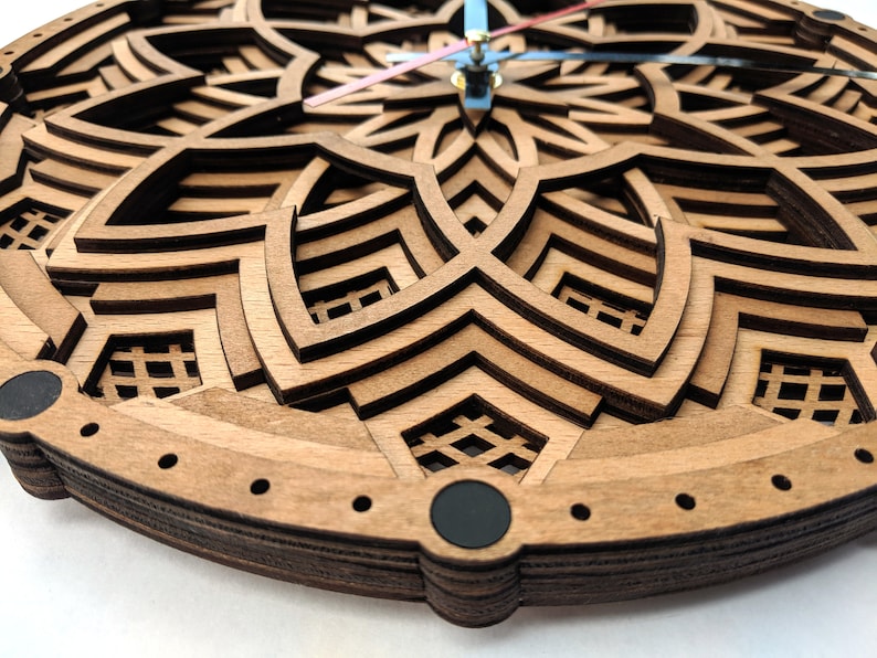 Op de afbeelding: Een houten wandklok met een gelaagd, geometrisch ontwerp. De klok heeft een lichtbruine kleur met ingewikkelde uitgesneden patronen. De wijzers van de klok zijn blauw en roze. De klok is een decoratief item.