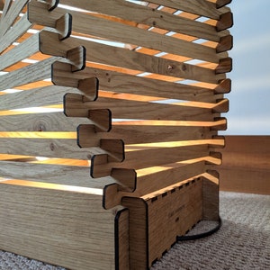 May include: A wooden lamp with a geometric design. The lamp is made of light brown wood and has a black finish. The lamp is lit and the light shines through the wood.