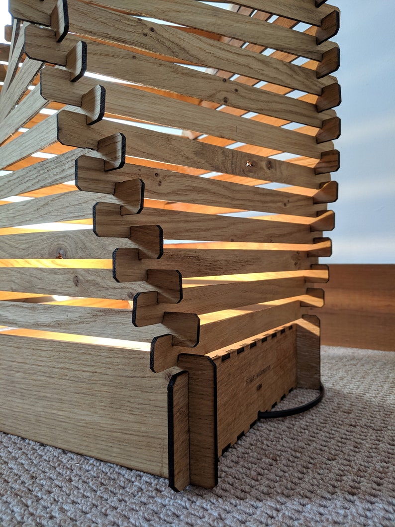 May include: A wooden lamp with a geometric design. The lamp is made of light brown wood and has a black finish. The lamp is lit and the light shines through the wood.