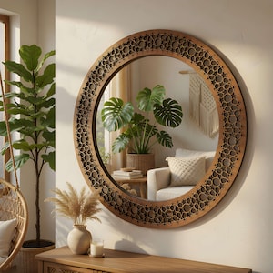 Peut inclure: Un grand miroir rond avec un cadre en bois décoratif orné d'un motif géométrique. Le miroir reflète un salon avec des plantes, un canapé blanc et une tenture murale en macramé. Le cadre est de couleur marron chaud.