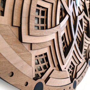 Op de afbeelding: Een gelaagde houten wandklok met een geometrisch ontwerp. De klok heeft meerdere lagen lichtbruin hout met donkerbruine accenten, waardoor een 3D-effect ontstaat. De wijzers zijn zilverkleurig. Het algehele ontwerp is modern en artistiek.