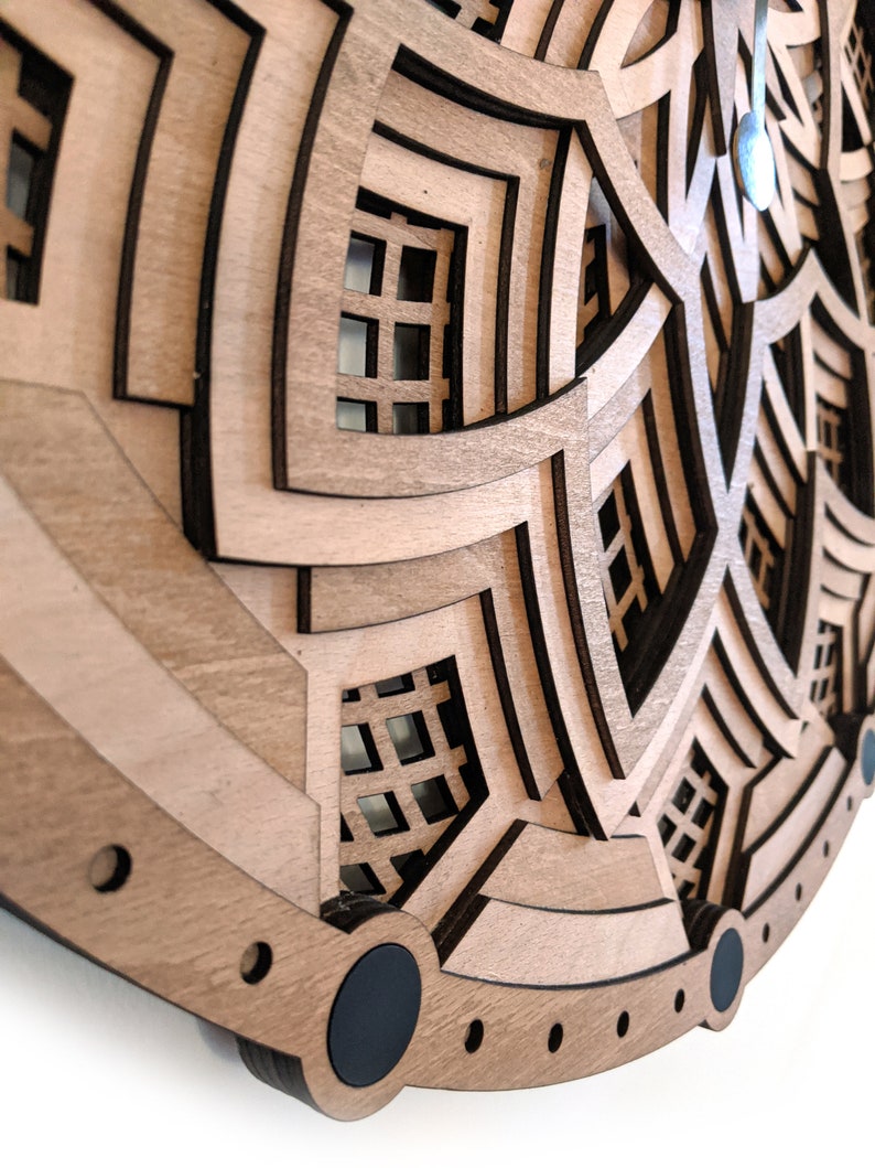 Op de afbeelding: Een gelaagde houten wandklok met een geometrisch ontwerp. De klok heeft meerdere lagen lichtbruin hout met donkerbruine accenten, waardoor een 3D-effect ontstaat. De wijzers zijn zilverkleurig. Het algehele ontwerp is modern en artistiek.