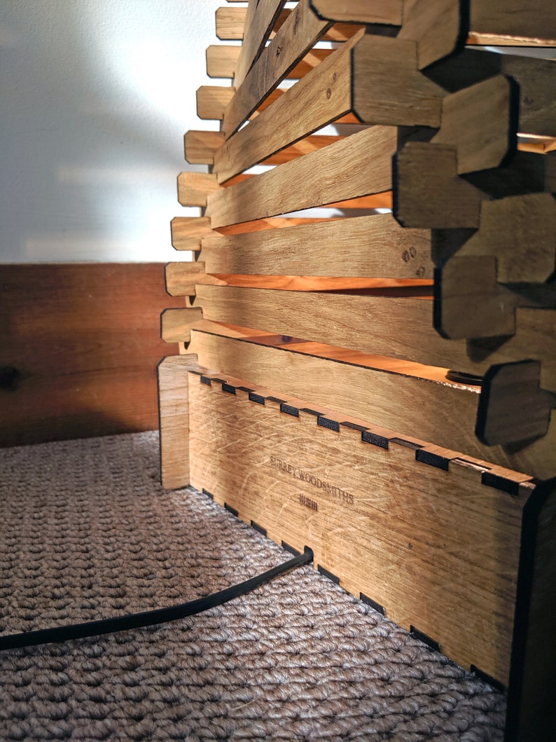 May include: A wooden lamp with a geometric design. The lamp is made of multiple pieces of wood that are stacked on top of each other. The lamp has a black cord and a label that reads "Surrey Woodsmiths".