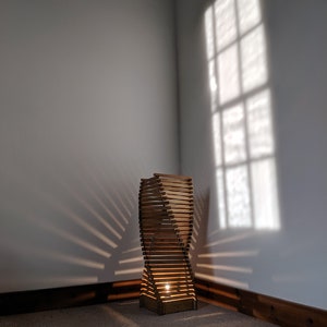 May include: A wooden floor lamp with a unique, twisted design. The lamp is made of thin wooden slats and has a warm, natural finish. The lamp is lit and casting a warm glow on the floor.