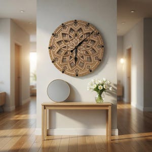 Op de afbeelding: Een grote, ronde houten wandklok met een gelaagd bloemenontwerp hangt boven een houten console. Een ronde spiegel en een vaas met witte bloemen staan op de tafel. De klok heeft zwarte wijzers en zwarte stippen.