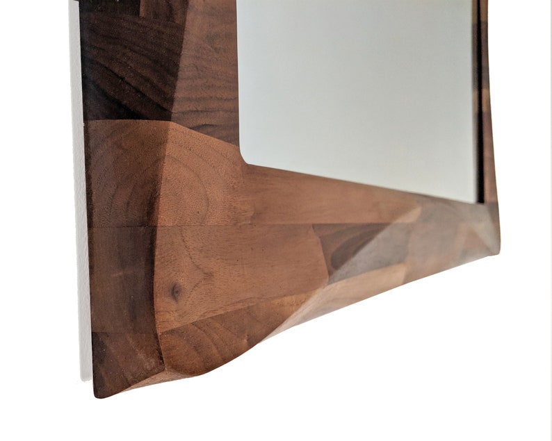 May include: A close-up of the bottom edge of a wooden mirror frame. The frame is made of multiple pieces of wood, each with a different grain pattern. The wood is a rich brown color.