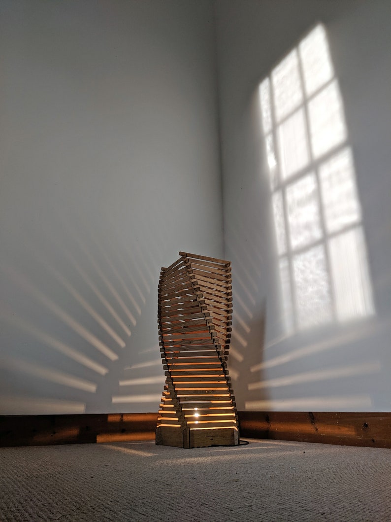 May include: A wooden floor lamp with a unique spiral design. The lamp is lit and casting a warm glow on the floor. The lamp is in a room with white walls and a window casting light through it.