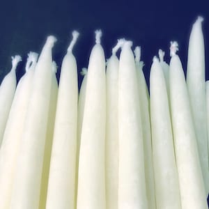 Puede incluir: Un montón de velas blancas de tipo taper, todas del mismo tamaño, están dispuestas en una pila. Las velas están todas apagadas.
