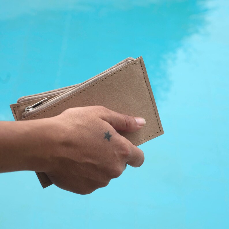 May include: A tan leather wallet with a zipper closure. The wallet is held in a hand with a small star tattoo.