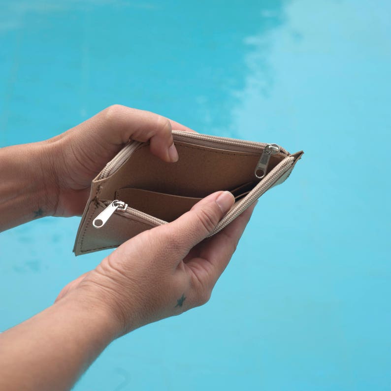 May include: A beige leather wallet is open and empty, held in a person's hands. The wallet has a zipper closure and is showing the inside lining.