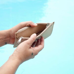 May include: An empty beige zippered pouch held open by two hands. The pouch is made of a textured material and has a silver zipper.