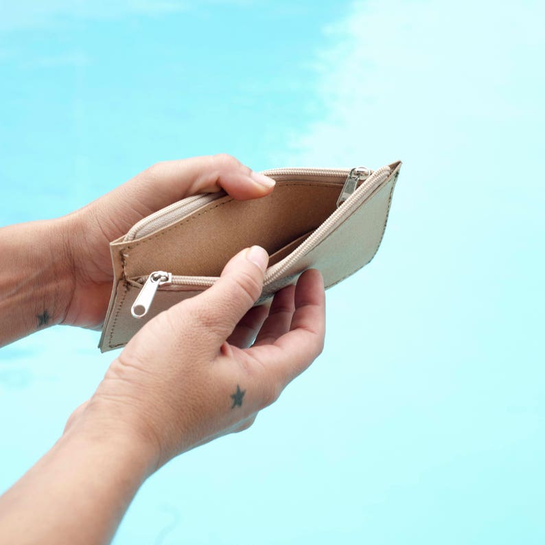 May include: An empty beige zippered pouch held open by two hands. The pouch is made of a textured material and has a silver zipper.