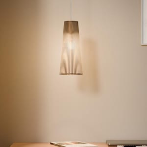 May include: A pendant light fixture with a brown, slatted shade hangs above a wooden desk. The light is on, illuminating the interior of the shade. A book and a laptop are on the desk. The light fixture is suspended from a thin white cord.