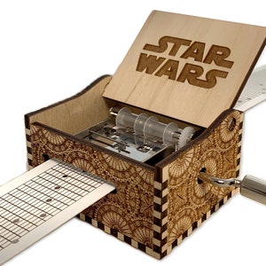 May include: A wooden music box with a Star Wars design on the lid. The music box is open and a paper music sheet is partially pulled out. The music box has a crank on the side.