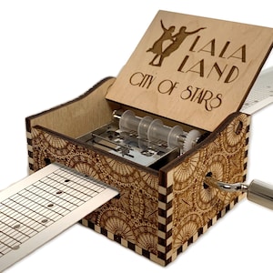 May include: A wooden music box with a hand-crank and a paper music sheet. The lid is open and features a laser-engraved design of two dancers with the text "La La Land City of Stars".