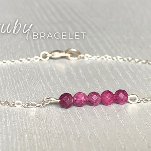 May include: A silver chain bracelet with a row of six pink ruby beads. The bracelet has a lobster clasp closure. "ruby bracelet" is written in the image.