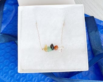 May include: A delicate gold-tone chain necklace featuring a horizontal arrangement of five small, raw, tumbled gemstone chips in shades of yellow, light green, black, reddish-brown, and clear, resting inside a white square gift box.