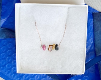 May include: A delicate necklace featuring a thin rose gold chain with five small, raw, multi-coloured gemstone chips arranged in a horizontal row. The stones include shades of pink, clear, brown, yellow, and black, suspended centrally on the chain.